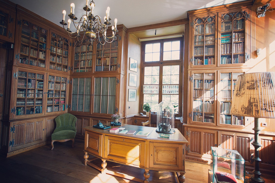 Die &raquo;Bibliothek&laquo;, einer der R&auml;ume im Museum auf Burg H&uuml;lshoff. Die W&auml;nde sind eingekleidet mit B&uuml;cherschr&auml;nken. In der Mitte des Raums steht ein gro&szlig;er Schreibtisch mit einem alten Mikroskop darauf. Au&szlig;erdem im Raum: ein Sessel, eine Lampe und ein Kronleuchter. Im Vordergrund steht eine kleine Vitrine mit Ohrringen aus dem Besitz Drostes.