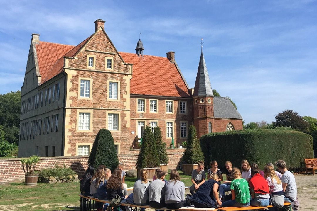 Eine Gruppe Jugendlicher Workshop-Teilnehmer*innen sitzt im Kreis aus dem Burghof. Im Hintergrund Burg H&uuml;lshoff.