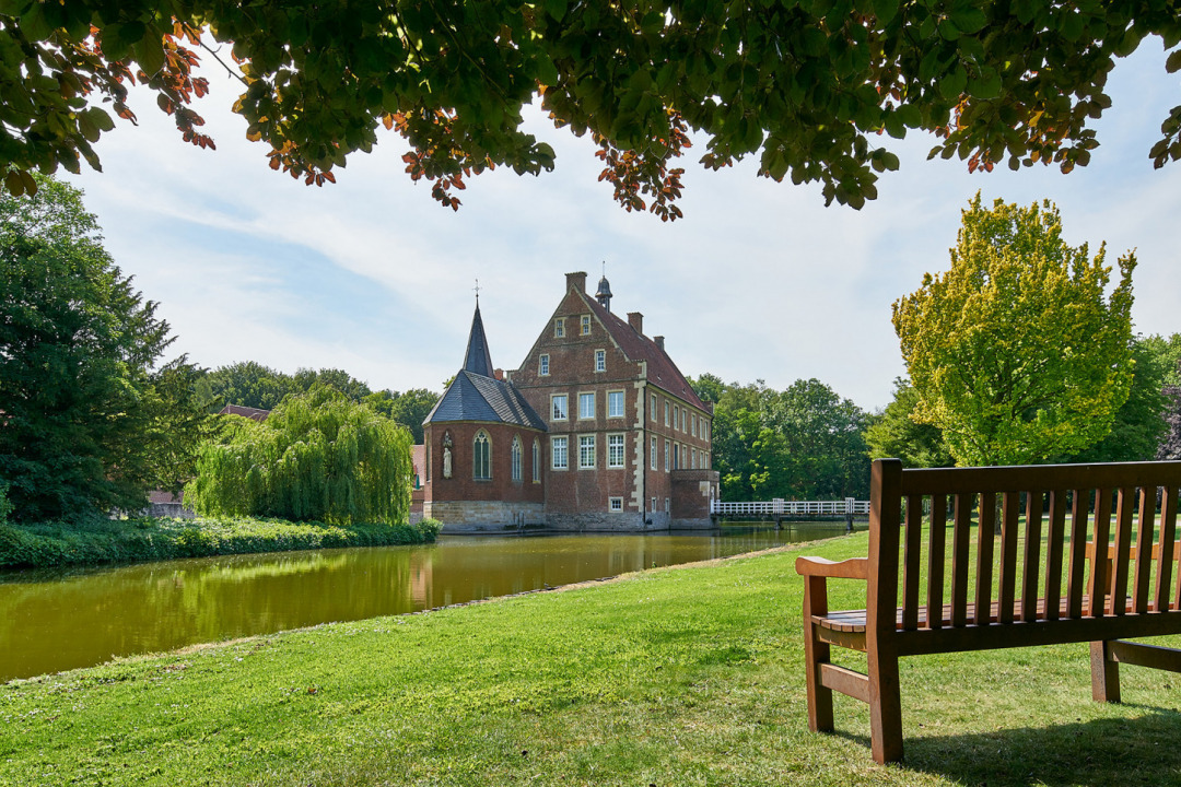 Bank im Park von Burg H&uuml;lshoff, ausgerichtet mit Blick auf Gr&auml;fte und Burg. Im Hintergrund B&uuml;sche und B&auml;ume.