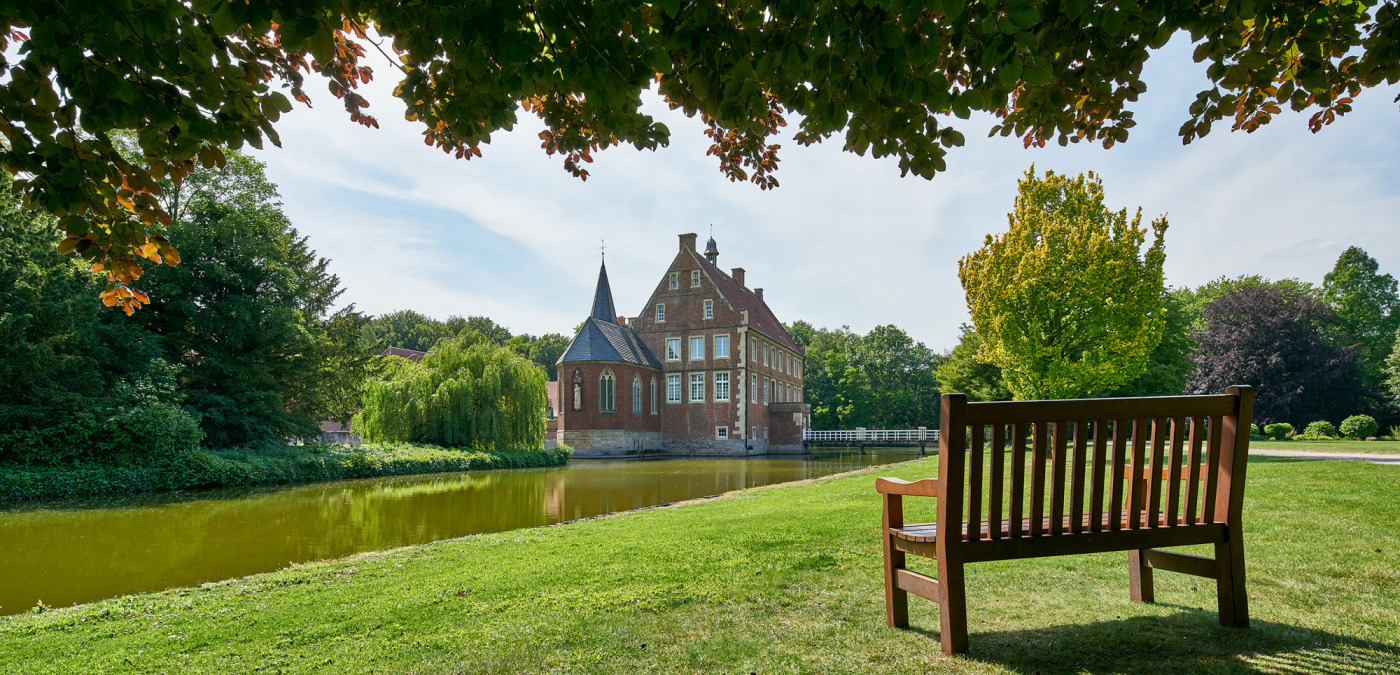 Bank im Park von Burg H&uuml;lshoff, ausgerichtet mit Blick auf Gr&auml;fte und Burg. Im Hintergrund B&uuml;sche und B&auml;ume.