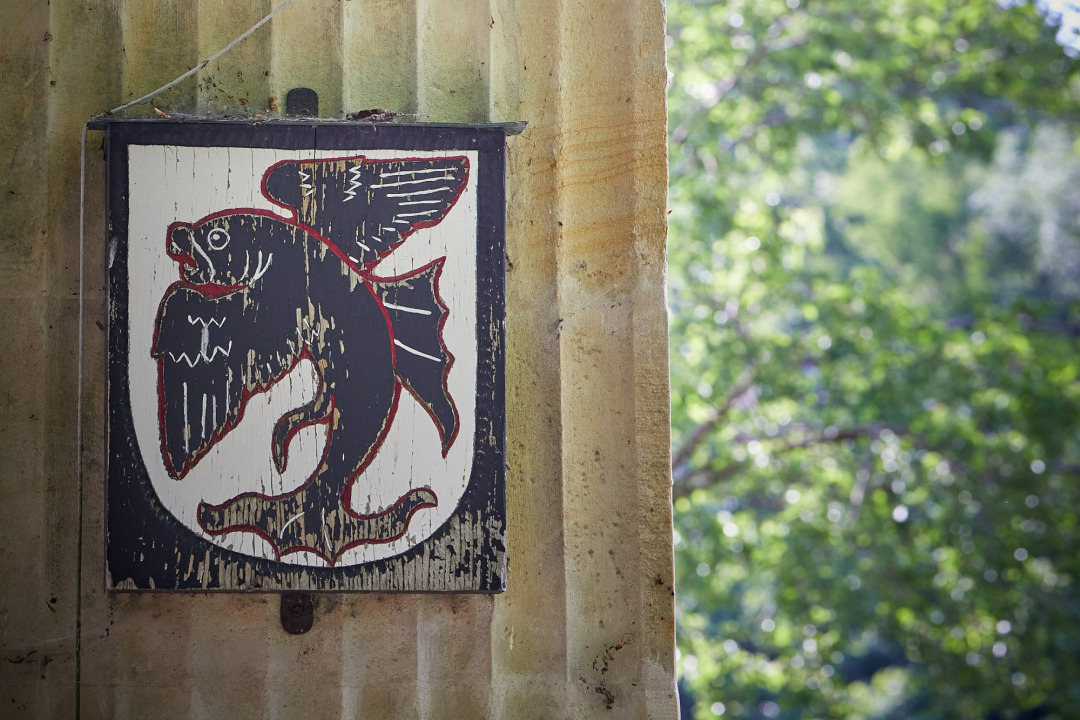 Wappen der Familie von Droste-H&uuml;lshoff an einem Sandstein-Torpfeiler auf einer Br&uuml;cke auf Burg H&uuml;lshoff. Das Familienwappen zeigt einen aufgerichteten, gefl&uuml;gelten Barsch, der sich zur linken Seite kr&uuml;mmt. An dem Pfeiler ist auch eine Glocke angebracht.