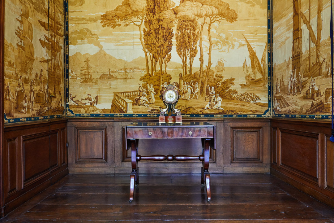 Foto, das im Inneren von Haus R&uuml;schhaus, im &raquo;Italienischen Zimmer&laquo;, aufgenommen wurde. Die Wand ist mit einer Tapete verkleidet, die noch aus der Zeit der Familie von Droste-H&uuml;lshoff stammt und Szenen im Hafen von Neapel zeigt. Au&szlig;erdem ein Tischchen mit einer alten Uhr.